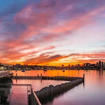 Coasterra restaurant overlooks calm water at sunset, with city skyline and colorful clouds in the background.