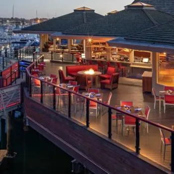 Outdoor restaurant with red chairs and wooden tables overlooks a marina filled with docked boats at dusk, with indoor and outdoor seating areas visible.