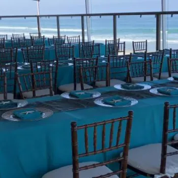 Rows of tables with turquoise tablecloths and set plates are arranged on a covered patio overlooking the ocean.