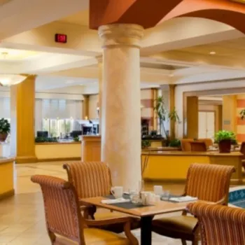 A spacious hotel dining area with tables, striped chairs, potted plants, and a central fountain, featuring columns and an open, well-lit layout.