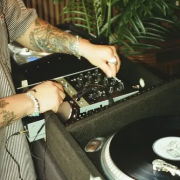 A person with tattooed arms adjusts knobs on a DJ mixer next to a turntable, with a plant in the background.