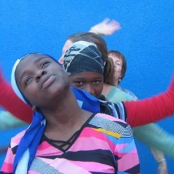 Four people stand in a line against a blue wall, with the front person tilting their head and others behind raising their arms, creating an illusion of multiple arms.