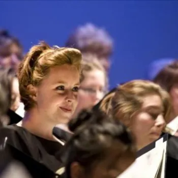 A group of choir singers holding sheet music, with one woman in the front row illuminated and looking forward, while others are focused on their music.