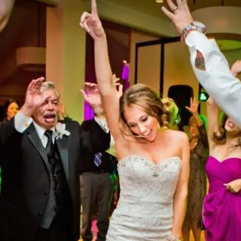 A bride in a white dress dances with guests in formal attire at a lively indoor wedding reception.