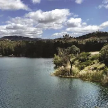 A calm lake surrounded by trees and hills under a partly cloudy sky, with two people in a small boat on the water.