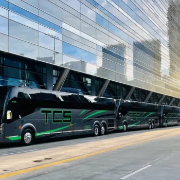 Several black and green TCS buses are parked in a row beside a modern glass building in an urban setting.