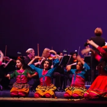 A group of dancers in colorful costumes perform on stage in front of an orchestra, with musicians playing string instruments in the background.