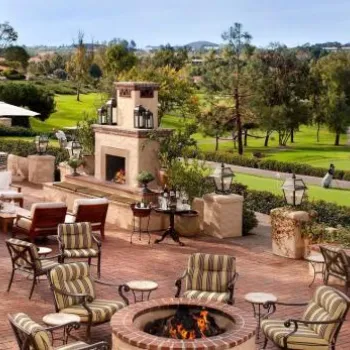 Outdoor patio with cushioned seating around a fire pit, stone fireplace, and dining area, overlooking a golf course and landscaped greenery.