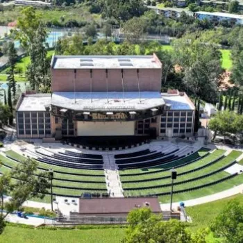 Aerial view of an outdoor amphitheater surrounded by trees, with curved seating facing a large stage, and buildings and greenery in the background.
