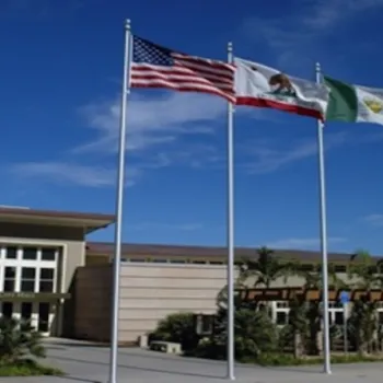 Three flags—the US flag, California state flag, and a green-and-white flag—fly in front of a modern building on a sunny day with blue sky and scattered clouds.