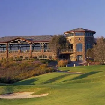 A large stone clubhouse with multiple windows and towers sits atop a hill, overlooking a green golf course with sand traps and scattered trees.