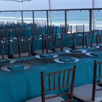 Rows of tables covered in teal tablecloths with place settings and brown chairs are arranged on a covered patio overlooking a beach and ocean.