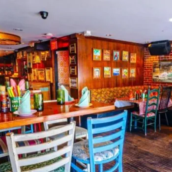 Colorful Cuban-themed restaurant interior with mismatched chairs, brick walls, framed photos, Cuban flag, and tables set with napkins and glasses.