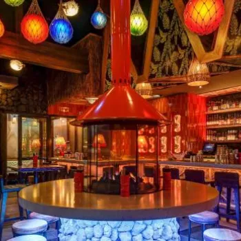 A dimly lit tiki bar with colorful hanging lanterns, a round central table with a fire feature, and a long bar lined with bottles and stools.