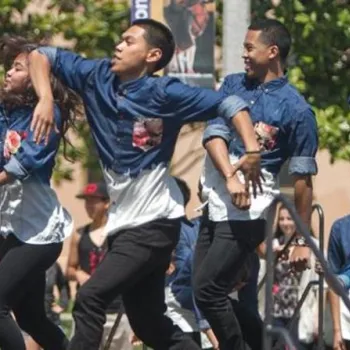 A group of five people in matching blue and white shirts perform a choreographed dance routine outdoors in front of an audience.