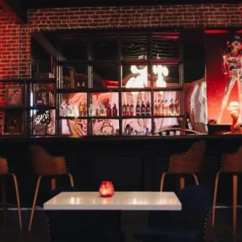 A dimly lit bar with high chairs, brick walls, modern art, and warm red lighting, featuring a well-stocked back shelf and candles on white tables.
