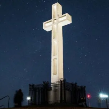 A large illuminated cross stands on a raised platform, surrounded by a fence, against a starry night sky.