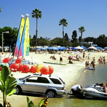 Crowds enjoy a sunny day at a sandy beach with colorful sailboats, tents, palm trees, and people preparing a jet ski near an SUV at the water’s edge.