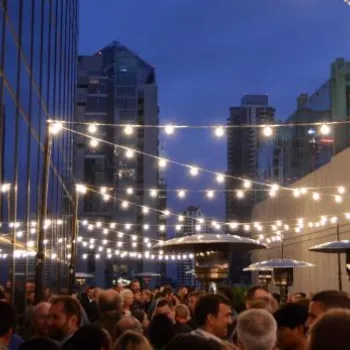 A large crowd gathers on a city rooftop patio in the evening, with string lights overhead and tall buildings in the background.