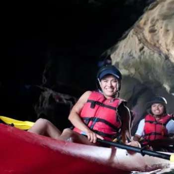 Three people wearing helmets and life jackets paddle a red kayak out of a dark cave into bright daylight.