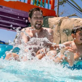 An adult and a child in swimsuits ride blue inner tubes and splash water in a pool at a water park, with rocks and plants in the background.