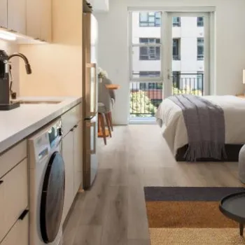 Modern studio apartment with a kitchen, washing machine, bed, and seating area, featuring large windows and contemporary decor.