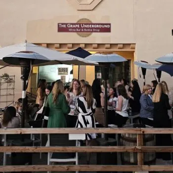 A group of people are gathered at outdoor tables under umbrellas at The Grape Underground wine bar during daylight.