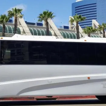 A large white shuttle bus is parked on a street in front of a modern glass building with palm trees and blue sky in the background.
