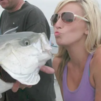 A woman in sunglasses leans in to pose as if kissing a large fish held by another person on a boat.
