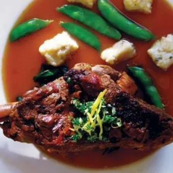 A cooked lamb shank with sauce, green snap peas, and small pieces of bread or dumplings on a white plate.