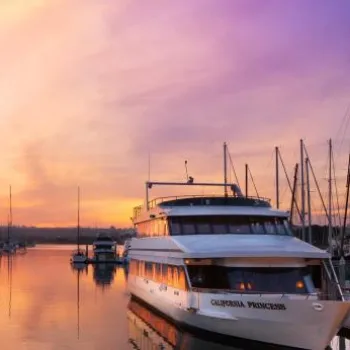 A docked yacht named "California Princess" surrounded by sailboats at a marina during a colorful sunset.