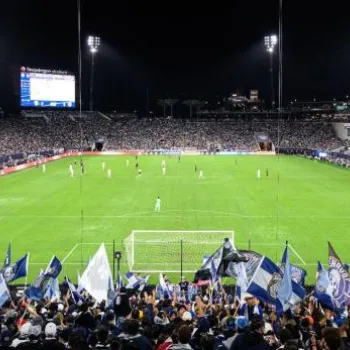 A large stadium filled with fans watching a soccer match at night; supporters in the foreground wave blue and white flags.