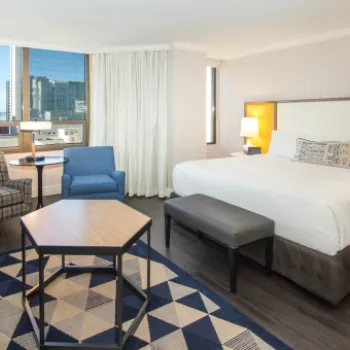 Modern hotel room with a king-size bed, seating area with chairs and a sofa, a large window with city view, and contemporary decor.