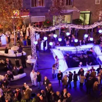 A crowd gathers at an outdoor event with illuminated booths and string lights at night, surrounded by trees and buildings.