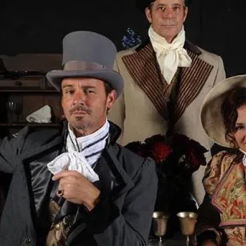 Three adults in Victorian-era costumes pose indoors; one man is seated wearing a gray top hat, another stands behind in a beige coat, and a woman in a bonnet sits next to them, smiling.