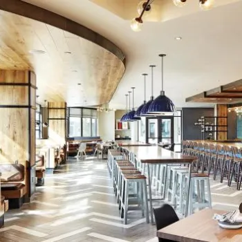 Modern restaurant interior with large windows, wooden accents, a long bar with blue tiles, high stools, booth seating, and pendant lighting.