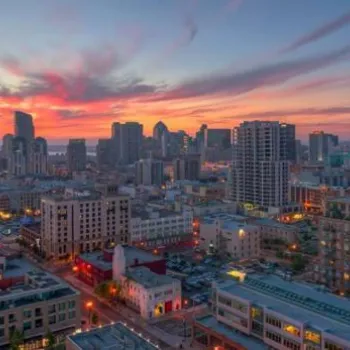Cityscape view at sunset with high-rise buildings, illuminated streets, and a colorful sky with clouds in the background.