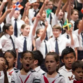 A large group of children in uniform sing together on stage, some holding small flags and raising their arms in enthusiasm.