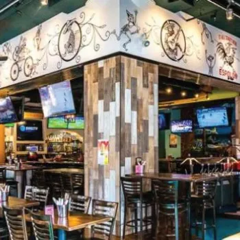 A colorful, modern bar and restaurant interior with wooden tables, high chairs, wall art, multiple TVs, and bright, decorative lighting.