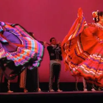 Two women in colorful traditional dresses perform a folkloric dance on stage, with musicians playing in the background against a pink backdrop.