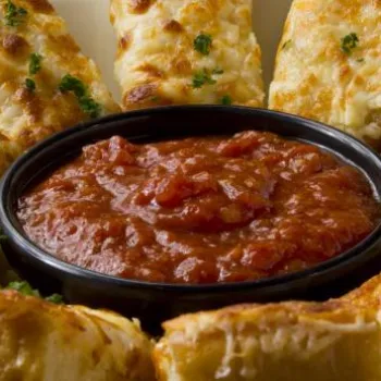 Slices of cheesy garlic bread arranged in a circle around a bowl of chunky tomato dipping sauce, garnished with chopped herbs.