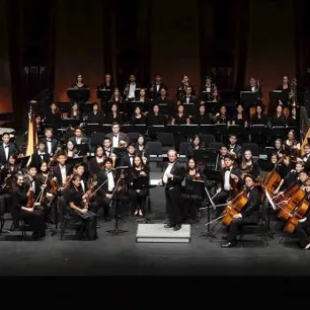 A full symphony orchestra is seated on stage in formal attire, with a conductor standing at the center in front of the musicians.