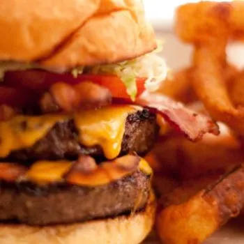 A cheeseburger with bacon, lettuce, and tomato on a bun, served with a side of crispy onion rings.