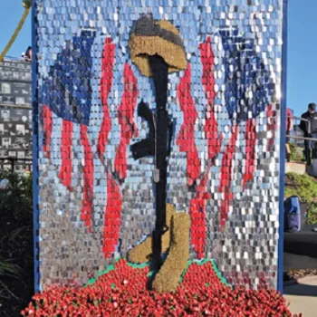 A memorial display featuring a helmet, rifle, and boots in front of an American flag mosaic, with red flowers at the base and people visible in the background.