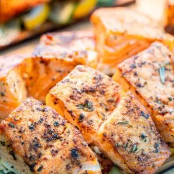Grilled salmon fillets seasoned with herbs, arranged on top of sliced zucchini and lemon, displayed on wooden serving boards.