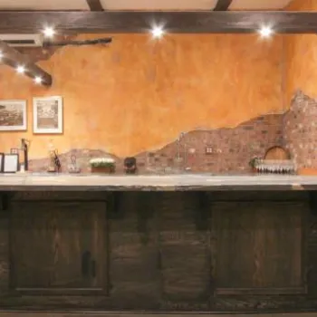 A rustic bar with a stone countertop, exposed brick walls, wine bottles, metal tanks on the left, and framed art hanging on a yellow textured wall.