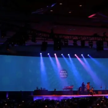 Wide stage setup with blue and purple lighting, musicians performing at center, and audience seated in a dimly lit large event hall.