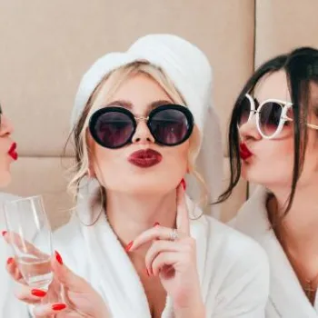 Three women in white bathrobes and towels on their heads wear sunglasses, hold champagne glasses, and pose playfully together, pursing their lips for the camera.