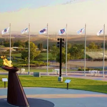 A large outdoor Olympic cauldron with a burning flame is surrounded by flagpoles displaying various national flags, with open fields and bleachers in the background.