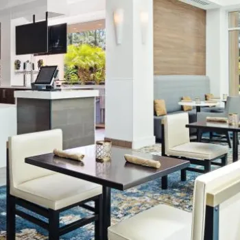 Bright, modern restaurant interior with white walls, blue patterned carpet, neatly set tables and chairs, bar area, and large windows letting in natural light.